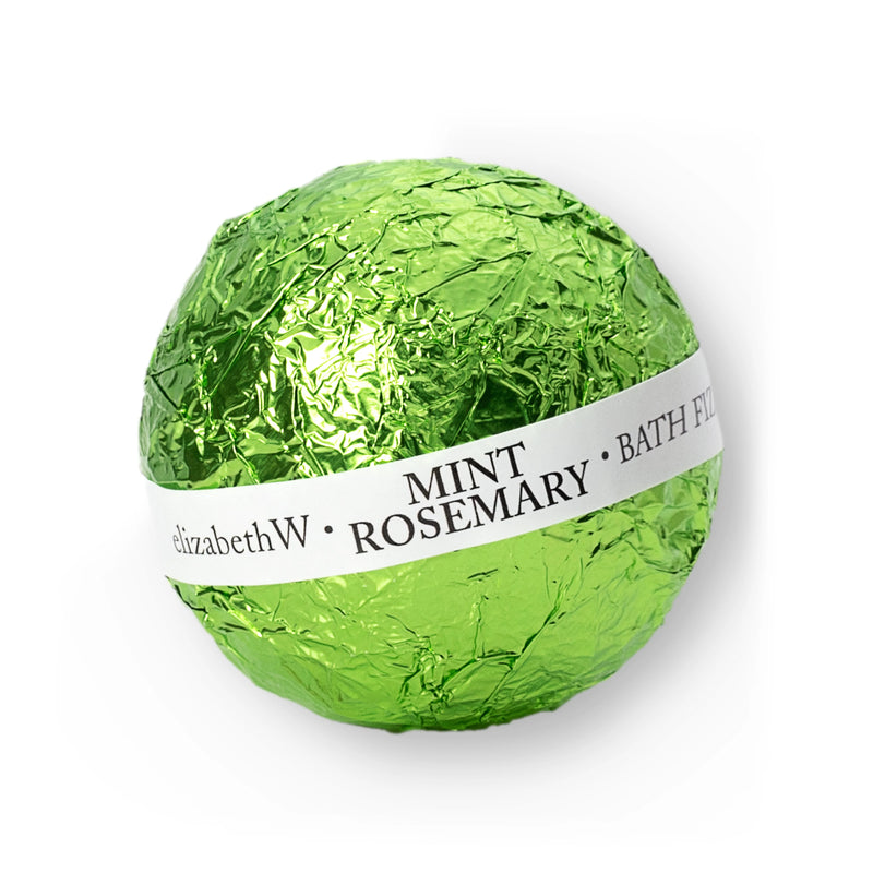 A mint and rosemary bath fizz wrapped in bright green foil with a white label displaying the product name, "elizabethW Purely Essential Mint Rosemary Fizz Ball." The spherical packaging suggests a refreshing, herbal bath.