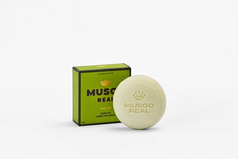 A bar of Claus Porto Musgo Real Classic Scent Shaving Soap by Claus Porto 1887 beside its green and black packaging, against a white background. The label on the soap matches the packaging design and promises a luxurious lather.