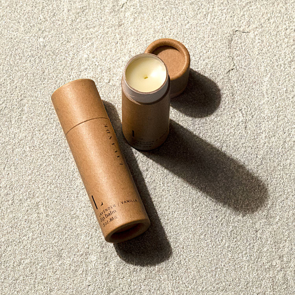 Two Lavande lip balms with natural ingredients in eco-friendly, wooden containers with caps removed, placed on a textured surface under sunlight casting sharp shadows.