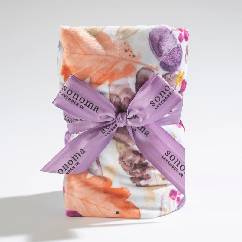 A rolled Sonoma Lavender Autumn Splendor Heat Wrap tied with a lavender ribbon labeled "Sonoma Lavender Co." on a white background, showcasing warm hues of orange and purple, and filled with flaxseed.