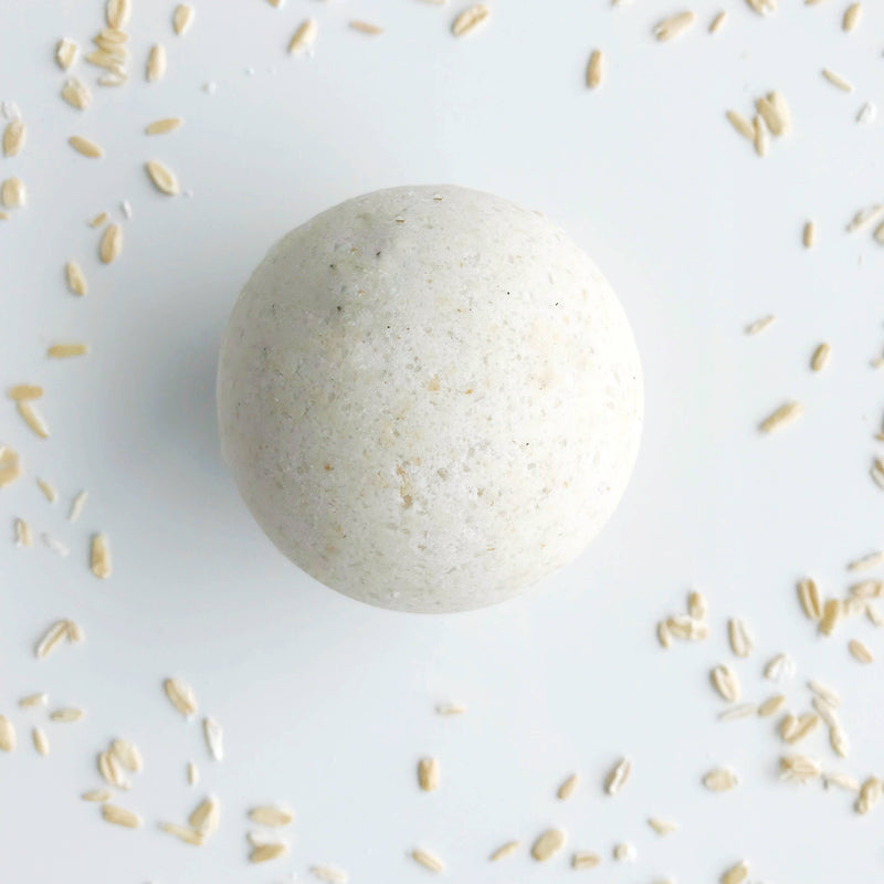 A single SOAK Bath Co. Oatmeal Milk and Honey bath bomb centered on a white background, surrounded by scattered rice grains.