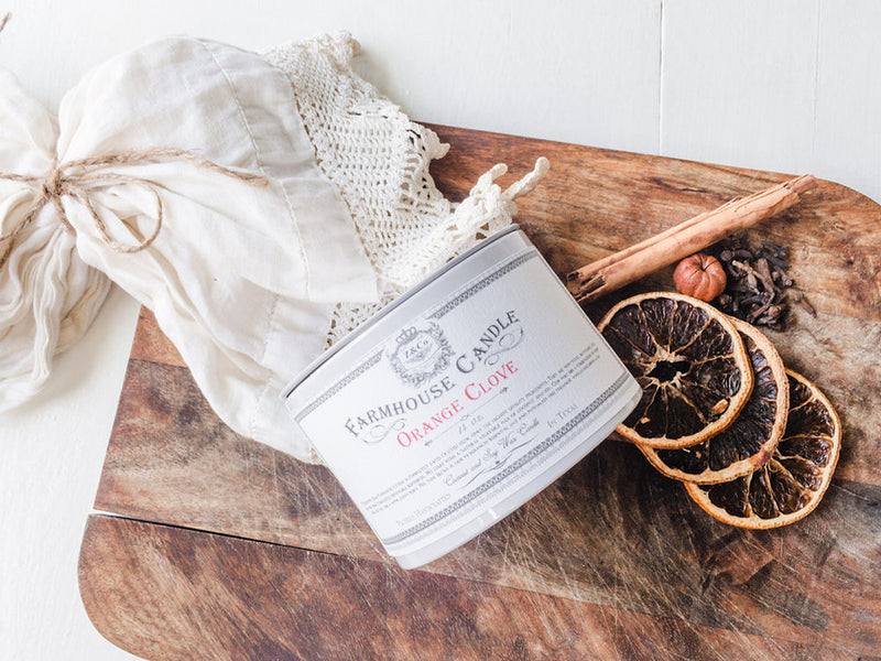A non-toxic candle labeled "Z&Co. Orange + Clove Farmhouse Candle" rests on a wooden board beside dried orange slices, nuts, and cinnamon sticks, with lace fabric in the background.