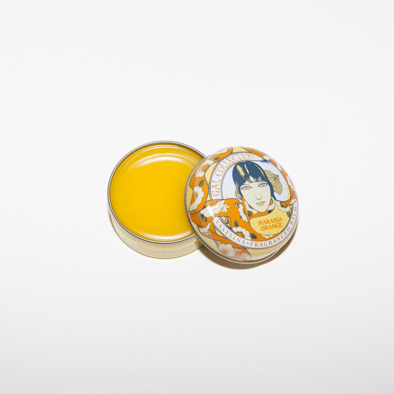 A small, open container of Perfumeria Gal Madrid Fragranced Lip Balm- Orange with a vividly colorful Art Nouveau Tin lid featuring illustrated artwork of a woman's face surrounded by text and decorative elements on a white background.