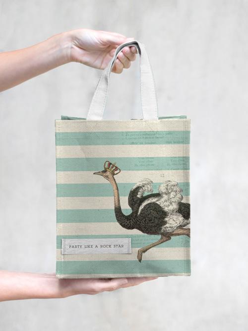 A hand holding a TokyoMilk tote bag with a vintage-style illustration of an ostrich and the phrase "party like a rock star" against a striped green background.