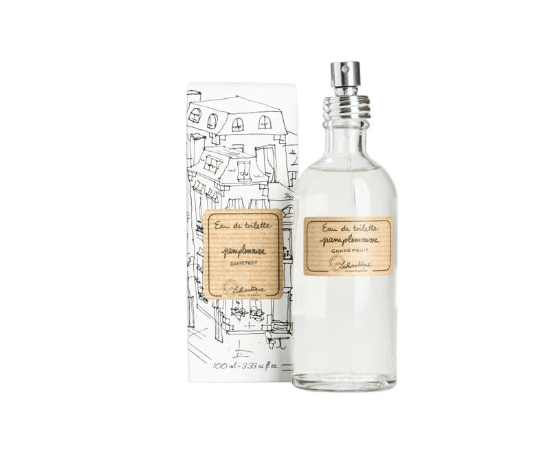 A clear glass Lothantique Pamplemousse Grapefruit Eau de Toilette bottle with a beige label next to its illustrated box, isolated on a white background. The label and box feature elegant black line drawings and script highlighting its grapefruit fragrance.