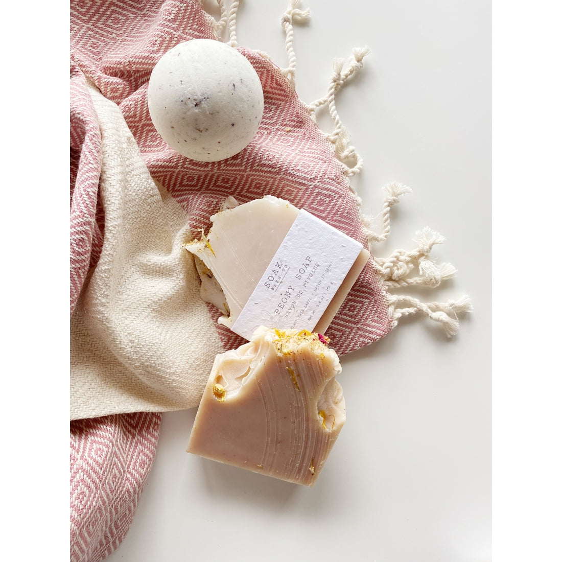 Handmade SOAK Bath Co. - Peony Soap Bars and a bath bomb displayed on a textured white surface alongside a pink and white patterned towel with tassel details.
