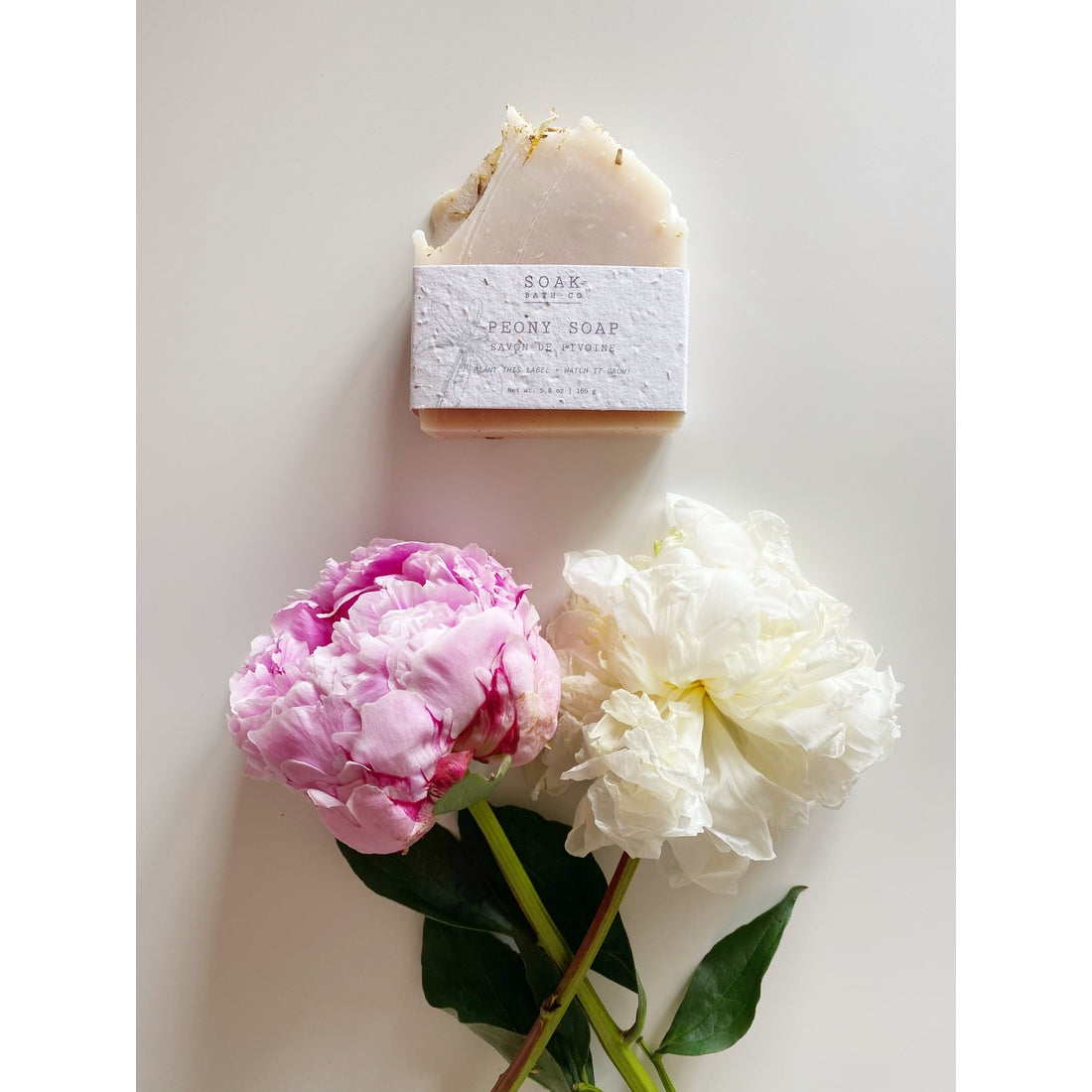 A bar of SOAK Bath Co. - Peony Soap Bar packaged in a white box with gold text, made from plantable seed paper, placed next to two lush peonies in pink and white, on a light beige surface