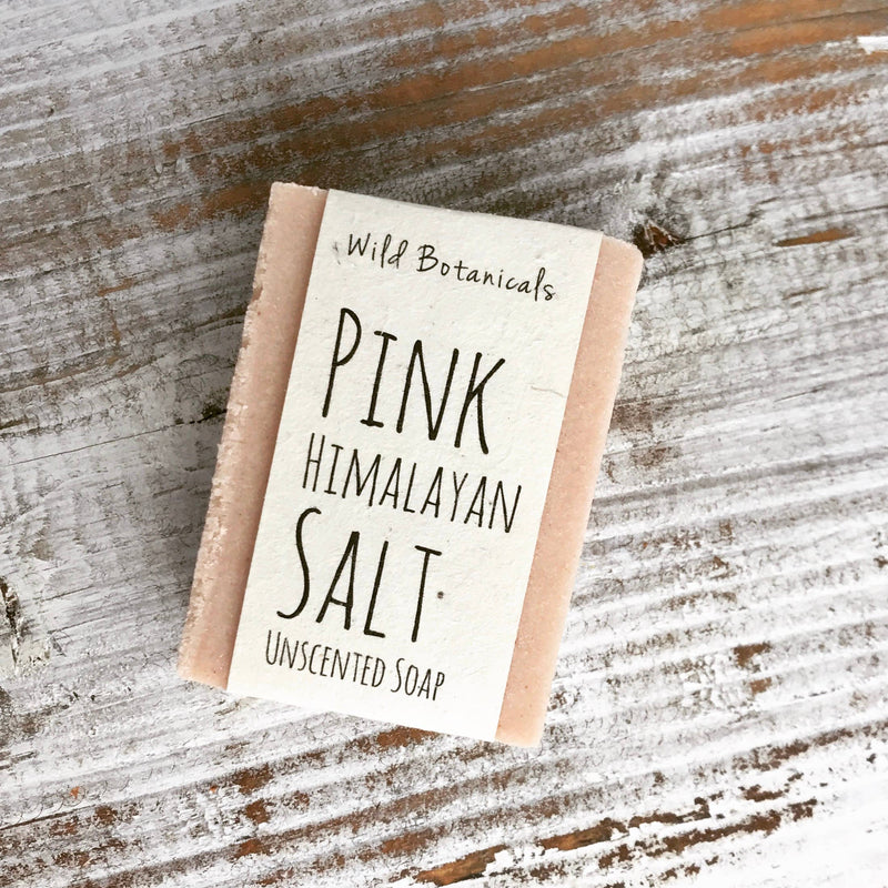 A bar of pink Himalayan salt exfoliating soap labeled "Wild Botanicals Pink Himalayan Salt Soap" placed on a rustic wooden surface.