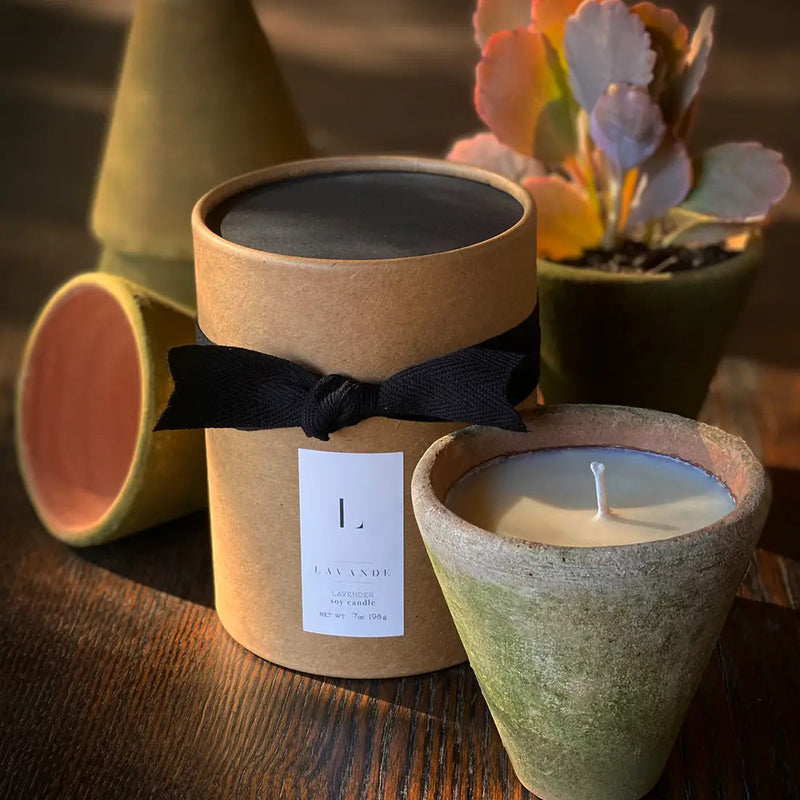 A photo of a Lavande lavender scented soy wax candle in a Lavande clay pot with a black ribbon tied around a cylindrical package, next to a potted succulent, on a wooden surface.