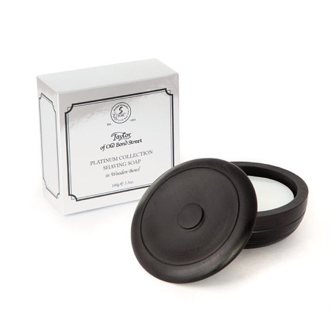 A round, black puck of Taylor of Old Bond Street Platinum Collection Shaving Soap in Wooden Bowl infused with woody oriental fragrance sits next to its open box labeled "Taylor of Old Bond Street Platinum Collection" on a white background.
