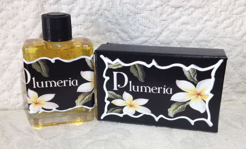 A bottle of Seventh Muse Fragrant Oil - Plumeria next to its box, both adorned with a white and yellow Plumeria flower design on a black background. The setting is against a textured white cloth.