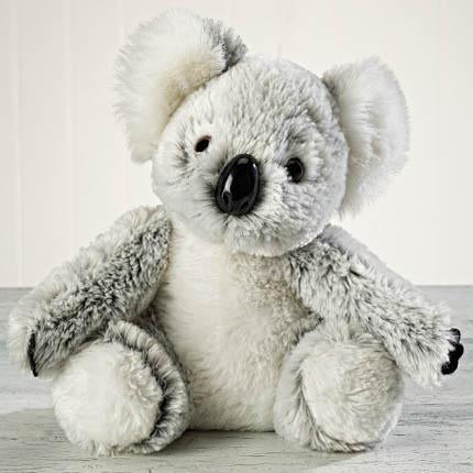 A Sonoma Lavender "Kaylee" plush toy sits on a white surface against a light wood panel background, with big black eyes, a prominent black nose, and fluffy grey ears.