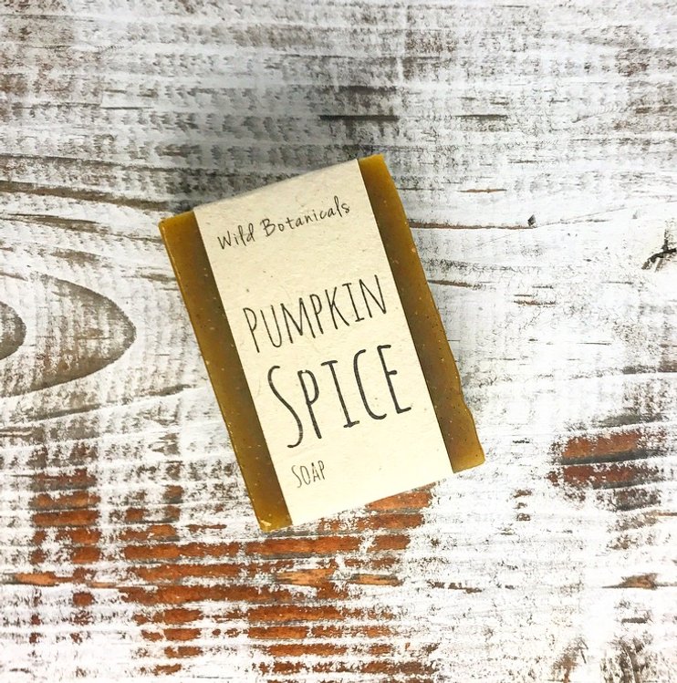 A bar of Wild Botanicals Pumpkin Spice Soap infused with organic shea butter lies on a rustic white and brown distressed wooden surface.