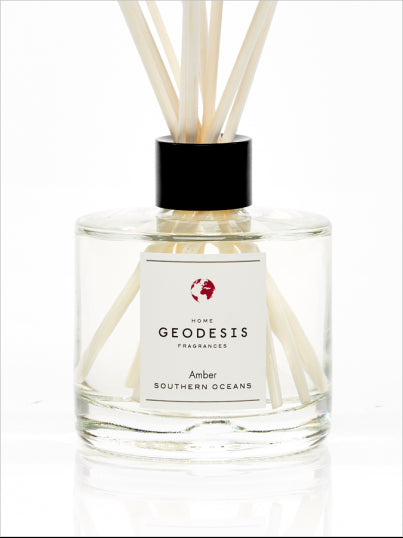 A clear glass diffuser bottle labeled "Geodesis Amber Reed Diffuser," featuring precious raw material with several reed sticks protruding from the top against a white background.