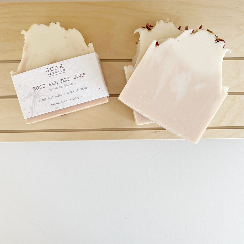 Two bars of "SOAK Bath Co. - Rosé All Day Soap Bar" placed on a light wooden surface, one with dried petals on top, next to its sustainable packaging. The soaps have a creamy color with textured edges.