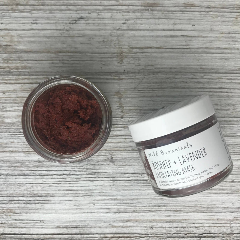 Top view of an open jar of Wild Botanicals Lavender & Rosehip Exfoliating Mask next to its closed container on a wooden surface. The container label includes product details.