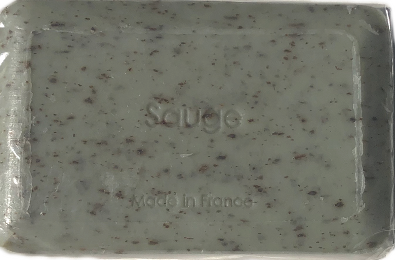 A close-up of a mint-green bar of La Lavande Sage Soap with dark specks, embossed with the word "savon" and "made in France" on its surface, contained in