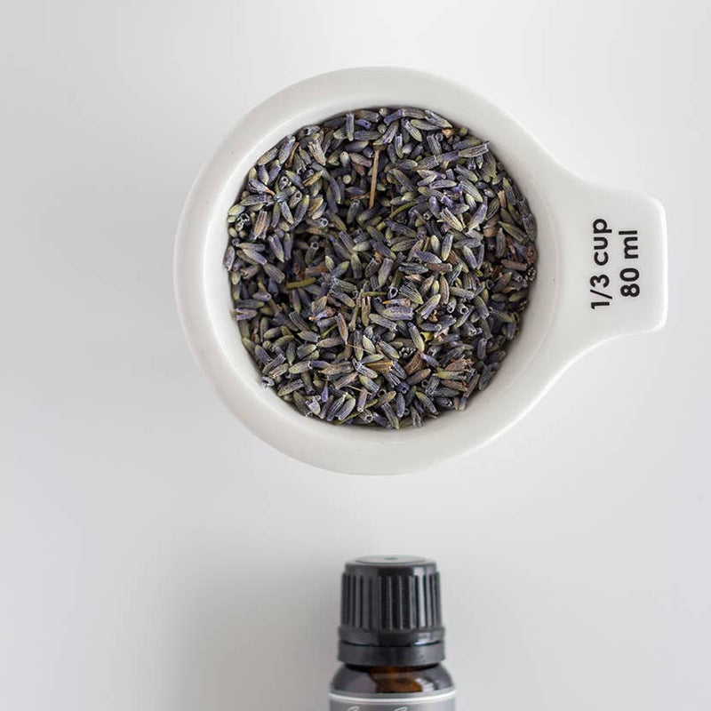 A top view of a 1/3 cup (80 ml) measuring cup filled with dried Sweet Streams Lavender Co. lavender buds, placed above a Sweet Streams Lavender Co. lavender essential oil bottle, against a plain white background.