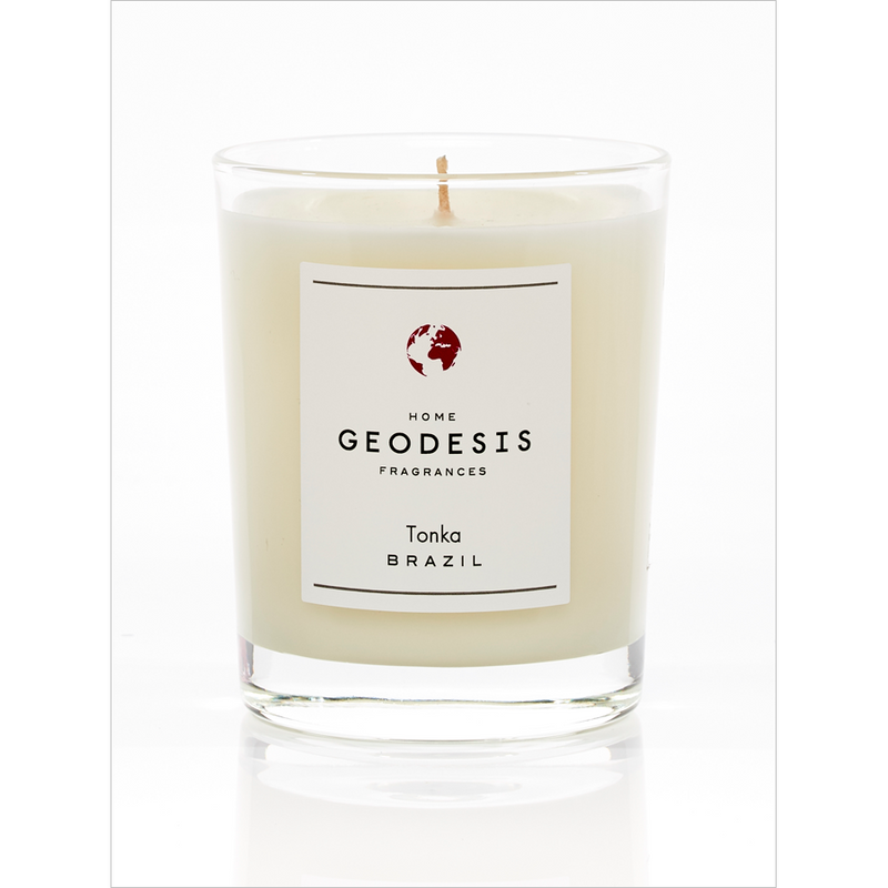 A large white candle in a clear glass container labeled "Geodesis" with "Tonka Brazil" and a burning time of 50 hours written underneath, featuring a small red and white.