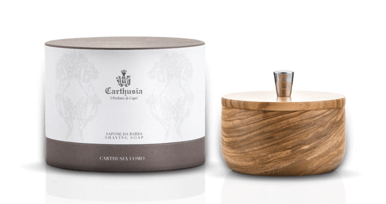 A cylindrical container with elegant gray packaging labeled "Carthusia Salone da Barba Olive Wood Shave Bowl With Soap" from the men's grooming collection, next to a round wooden lid with a metallic knob, isolated on a white background. Brand Name: Carthusia I Profumi de Capri