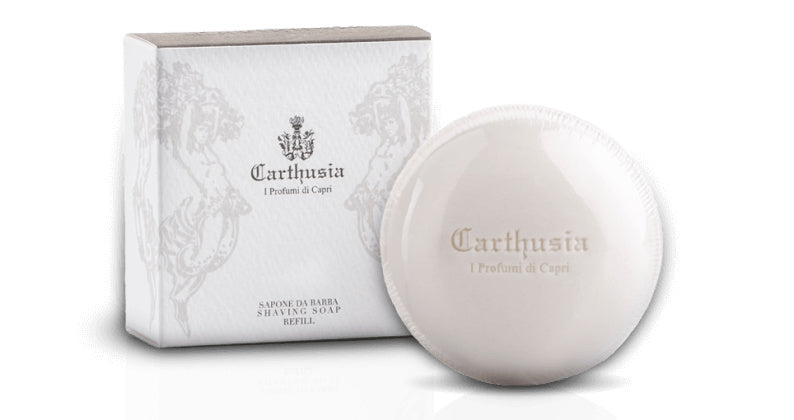 A round white men's Carthusia I Profumi de Capri shaving soap next to its gray and white floral-designed box labeled "Carthusia Uomo." The Carthusia Salone da Barba Shave Soap Refill is imprinted with the brand name.