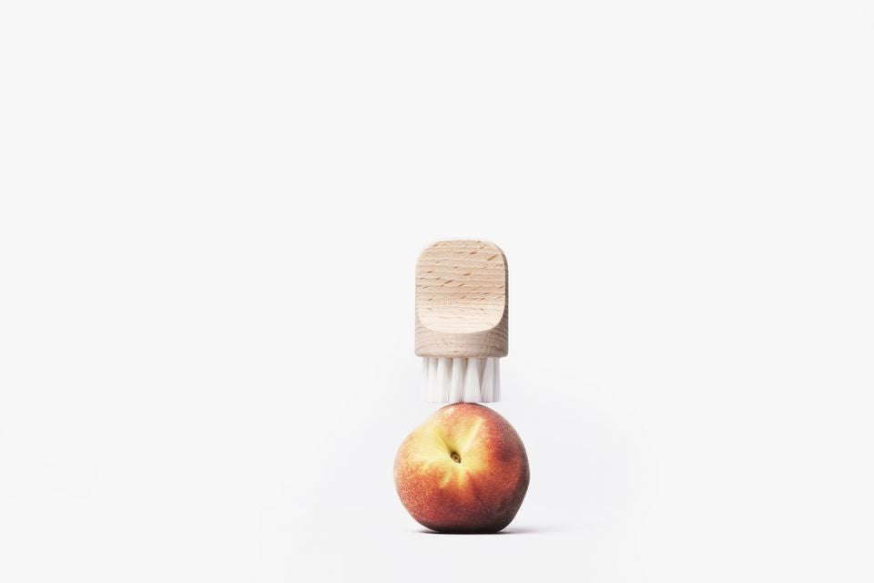A minimalist composition featuring an Andrée Jardin "Canot" Vegetable Brush Soft mounted on a ripe peach, set against a stark white background.