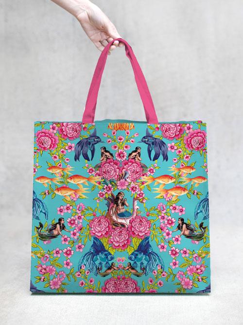 A person holds a TokyoMilk Tote Bag - Song of the Siren Market Tote by Margot Elena, with a vibrant floral and peacock design, set against a plain gray background.