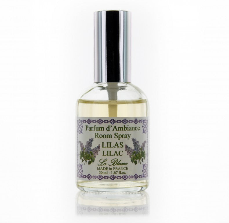 A clear glass bottle of "Le Blanc Lilac Room Spray" room spray with a silver cap, displaying a purple and green label with floral accents, on a seamless white background.