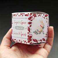 A hand holding a cylindrical Dot & Lil Sugar + Spice Sparkling Milk Bath Cube packaging labeled with illustrations of a rabbit, foliage, and berries in red and white tones.