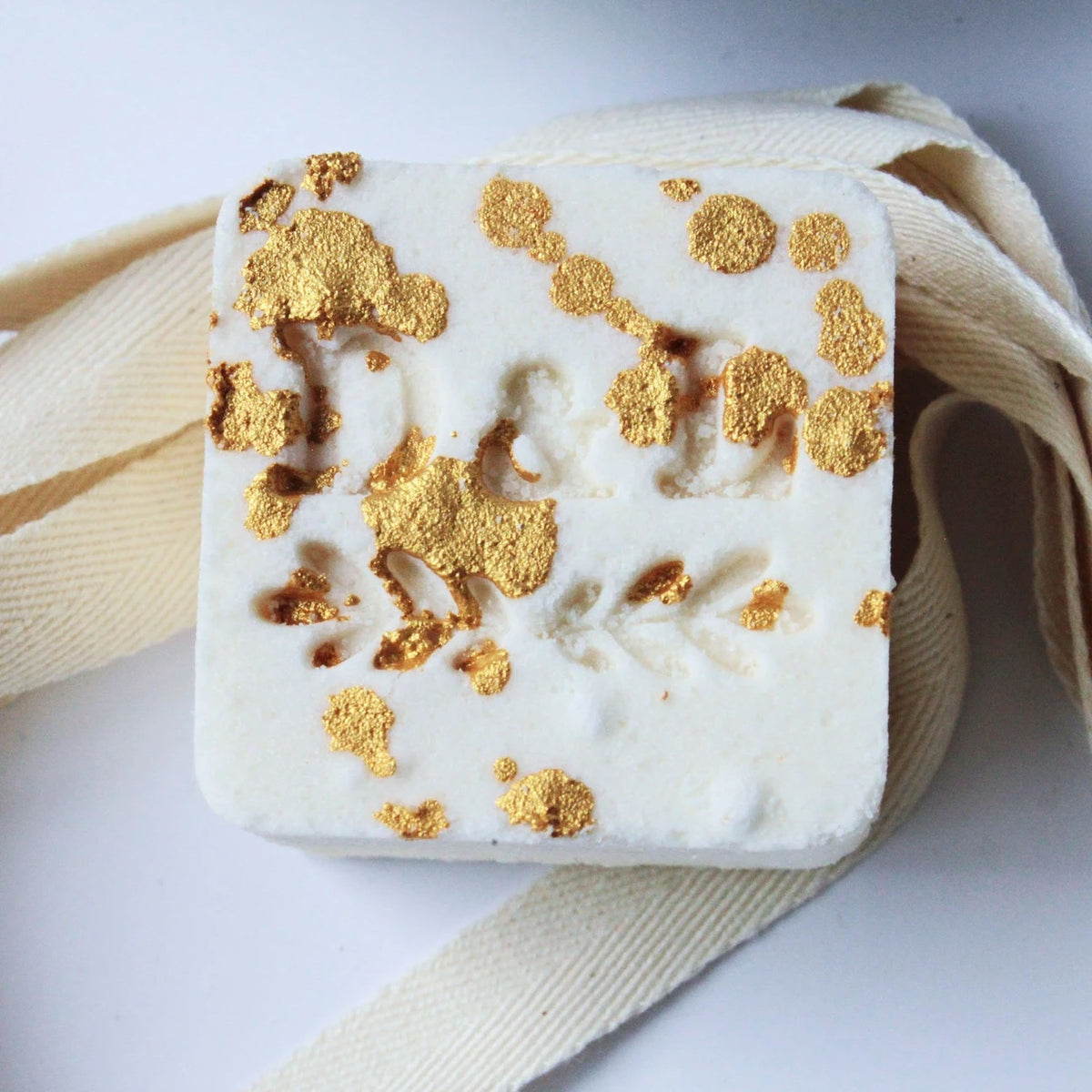 A square, handmade Dot & Lil Sugar + Spice Sparkling Milk Bath Cube with an embossed floral design, featuring patches of gold leaf scattered across its surface. It rests on a white background with a ribbon beside it.