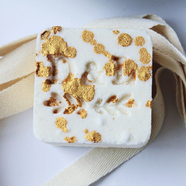A square, handmade Dot & Lil Sugar + Spice Sparkling Milk Bath Cube with an embossed floral design, featuring patches of gold leaf scattered across its surface. It rests on a white background with a ribbon beside it.