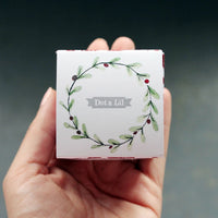 A person holds a Dot & Lil Sugar + Spice Sparkling Milk Bath Cube, with a delicate green wreath design and the text "Sugar & Spice" printed in the center, set against a blurry grey background.