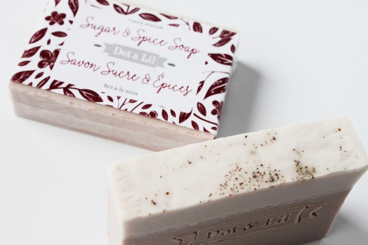 A bar of Dot & Lil Sugar + Spice Bar Soap with visible specks beside its packaging featuring elegant floral motifs and stylish typography. The soap and box rest on a white background.