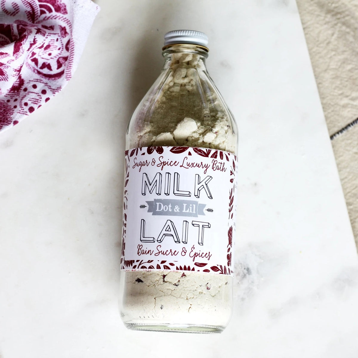 A clear glass bottle of Dot & Lil Sugar + Spice Milk Bath labeled "milk dot a lil" on a marble surface, with a decorative theme featuring red and white text and infused with moisturizing whole milk powder.