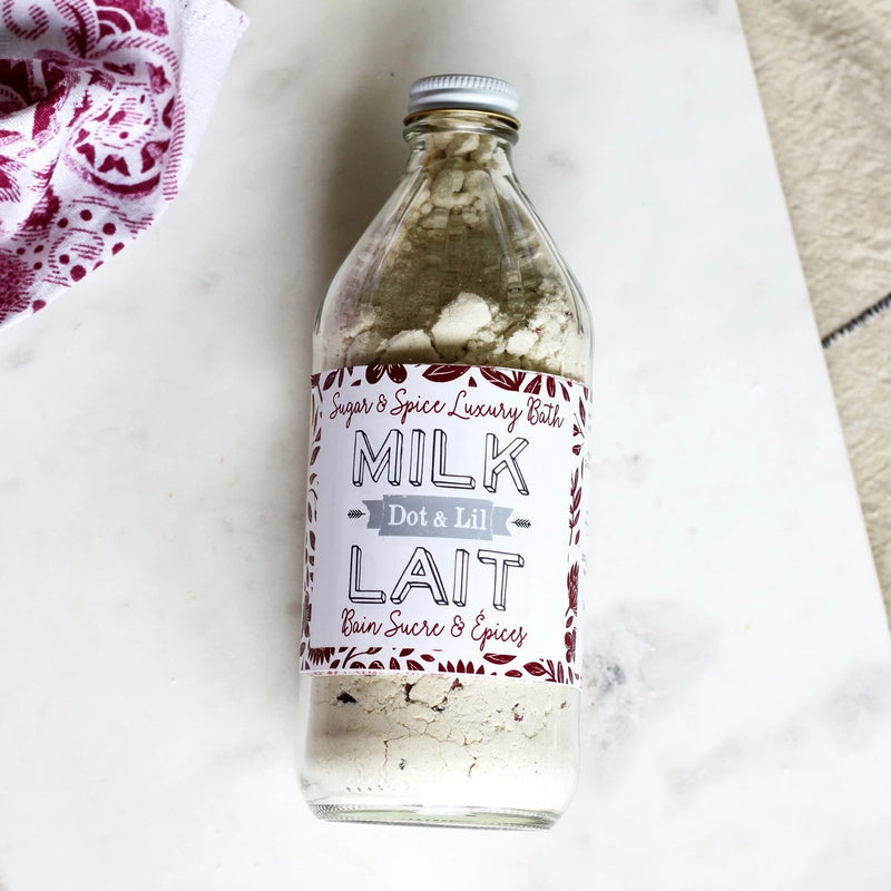 A clear glass bottle of Dot & Lil Sugar + Spice Milk Bath labeled "milk dot a lil" on a marble surface, with a decorative theme featuring red and white text and infused with moisturizing whole milk powder.