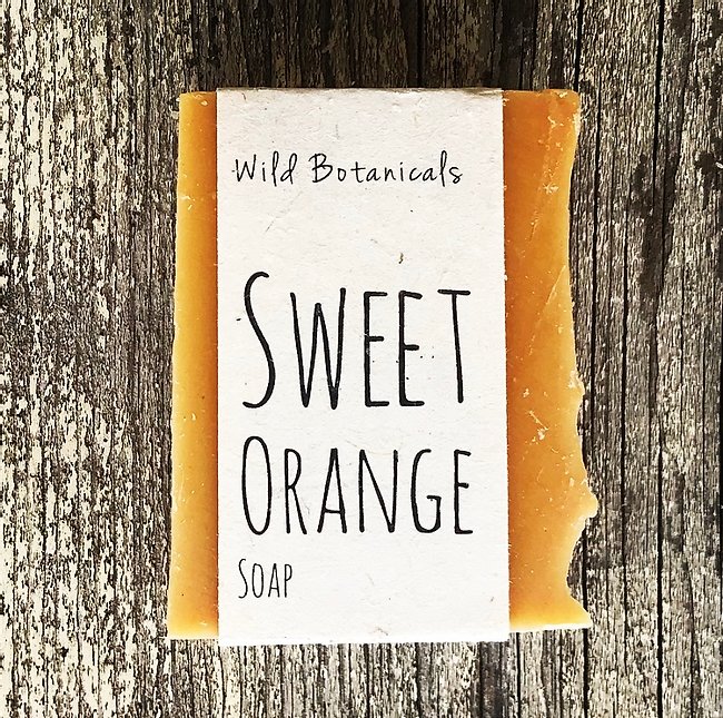 A bar of Wild Botanicals Sweet Orange Soap infused with essential oils from wild botanicals rests on a rustic wooden surface, with its label prominently displayed in a simple, elegant font.
