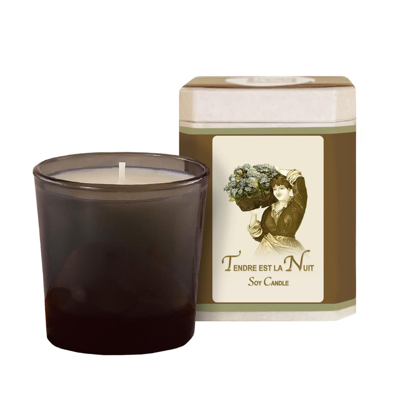 A dark brown glass candle next to a beige La Bouquetiere Tendre est la Nuit Soy Candle jar featuring a vintage illustration of a woman holding a bouquet and scented with Ylang Ylang.