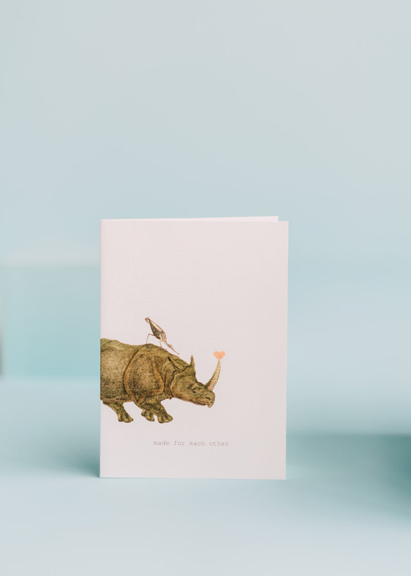 A TokyoMilk Greeting Card - Made For Each Other featuring an illustration of a rhinoceros with a bird on its back, holding a heart balloon. The text "made for each other" is at the bottom. The card is created by Margot Elena.