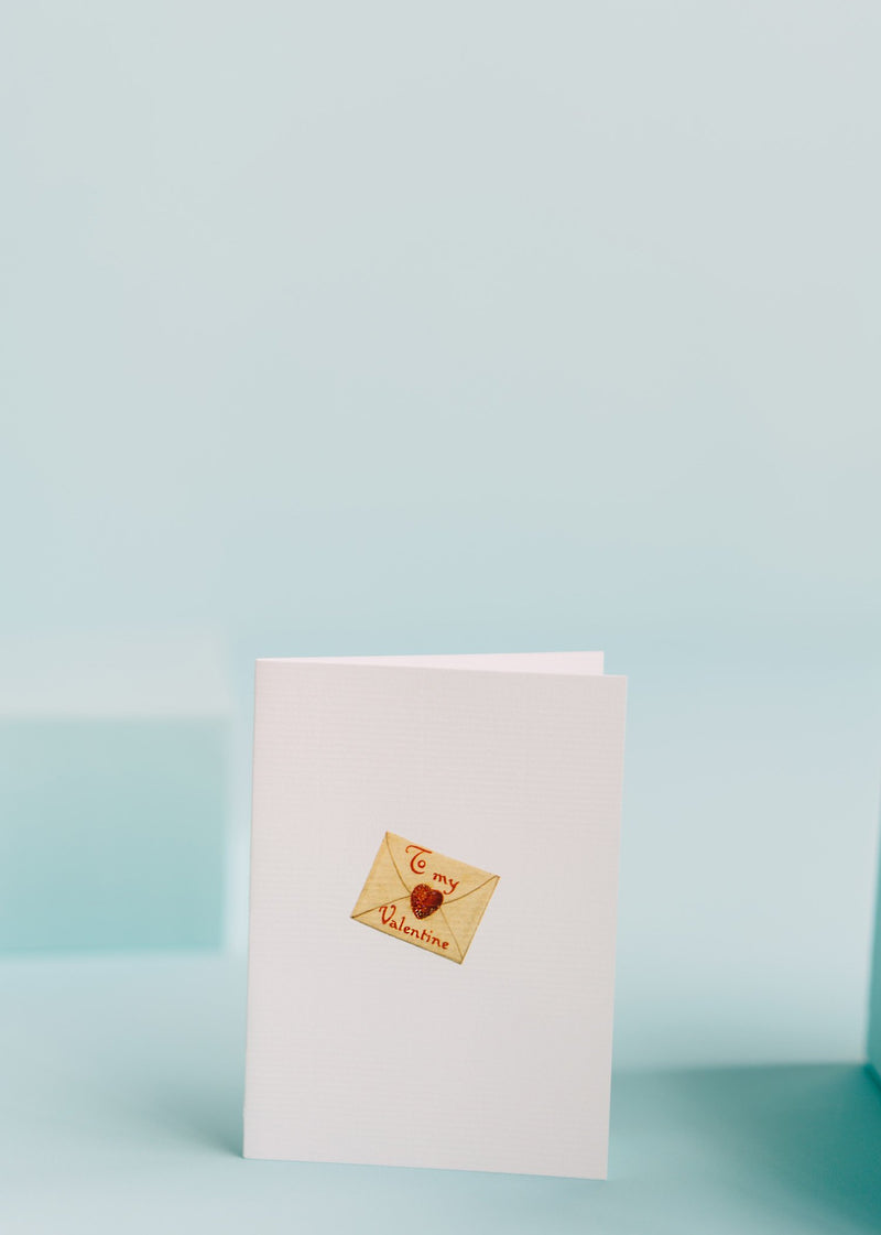 A TokyoMilk Greeting Card - To My Valentine stands open on a pale blue background. The card features a small, heart-shaped design with hand-glittered accents and the text "To my Valentine" in cursive script by Margot Elena.
