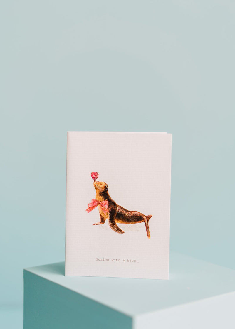 A TokyoMilk greeting card featuring a playful illustration of a seal balancing a red heart on its nose, with the caption "sealed with a kiss," set against a soft blue background and hand-glittered accents. Designed by Margot Elena.