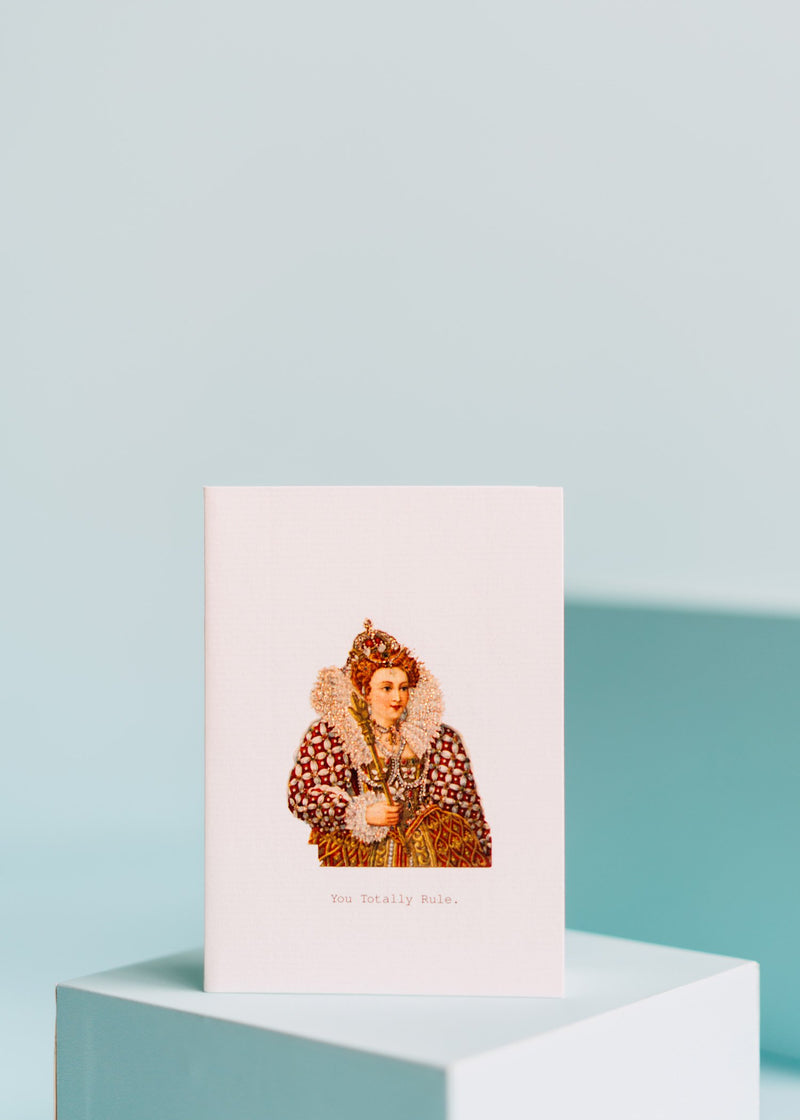 A Margot Elena greeting card featuring a hand-glittered illustration of a regal figure in historical dress with the text "you totally rule" below, displayed on a white pedestal against a soft blue background.