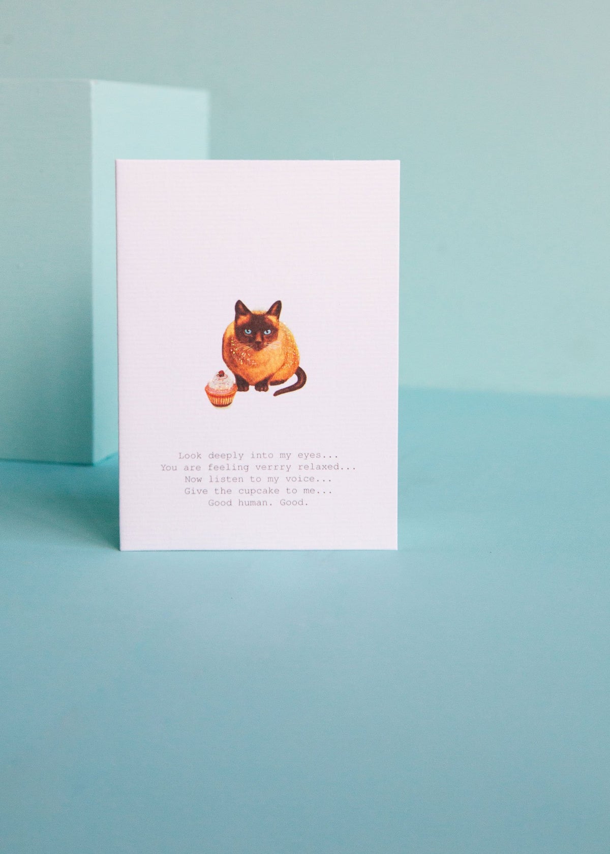A Margot Elena TokyoMilk Greeting Card featuring an illustration of a cat with hypnotic eyes and a caption that reads, "look deeply into my eyes... you are feeling very sleepyminded... give the cupcake to me