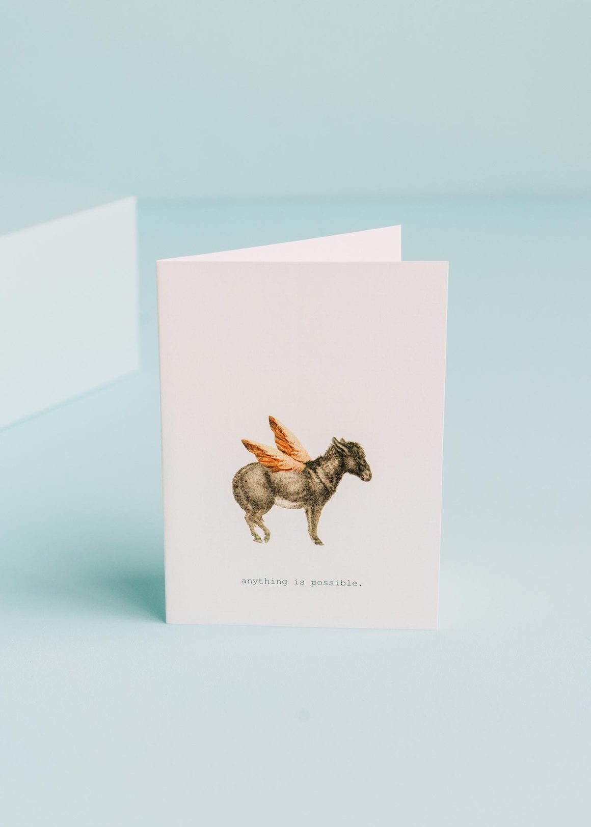 A TokyoMilk Greeting Card - Anything is Possible with a whimsical drawing of a winged donkey stands open on a light blue background, hand-glittered accents sparkling around the edges, and the text "anything is possible." Created by Margot Elena.