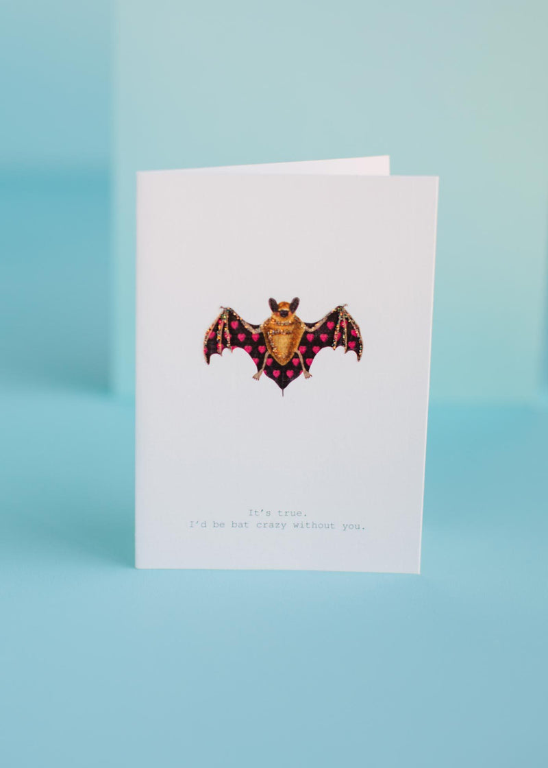 A TokyoMilk Greeting Card - Bat Crazy Without You by Margot Elena, with a colorful bat illustration, hand-glittered accents, and the text "it's true. i'd be bat crazy without you." stands against a light blue background.