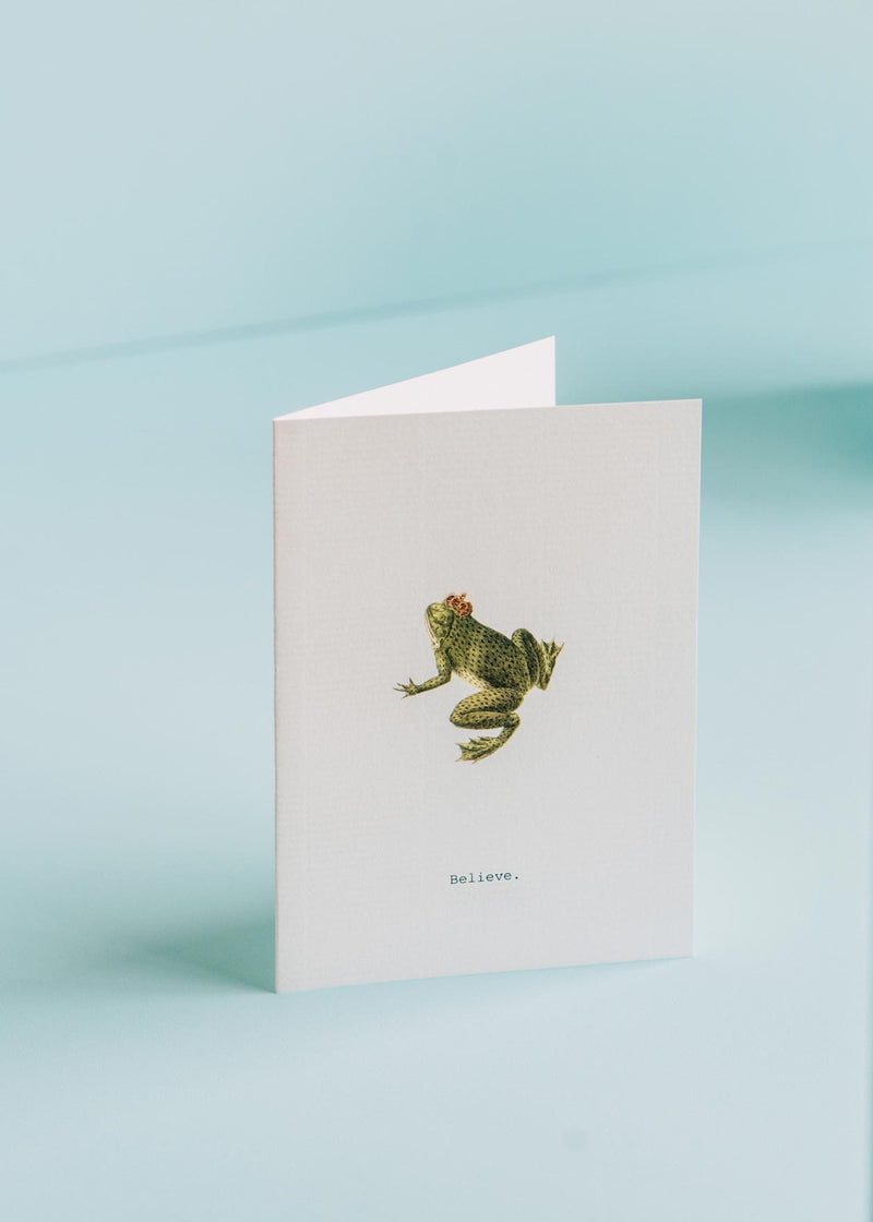 A TokyoMilk greeting card featuring an illustration of a frog on a light blue background. The word "believe" is printed at the bottom of the card on laid paper. Brand: Margot Elena