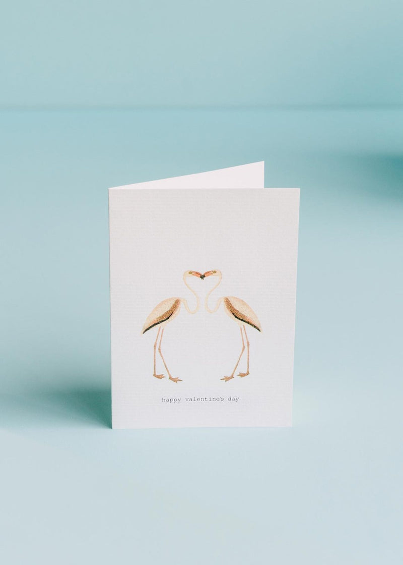 A TokyoMilk greeting card featuring two flamingos forming a heart shape with their necks and heads, with the phrase "happy valentine's day" printed below, against a soft blue background, and hand-gl