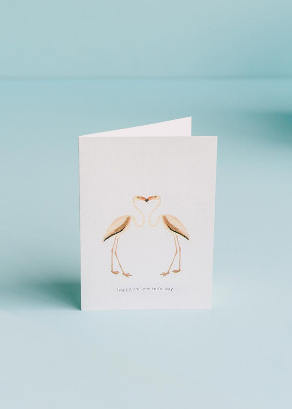 A TokyoMilk greeting card featuring two flamingos forming a heart shape with their necks and heads, with the phrase "happy valentine's day" printed below, against a soft blue background, and hand-gl