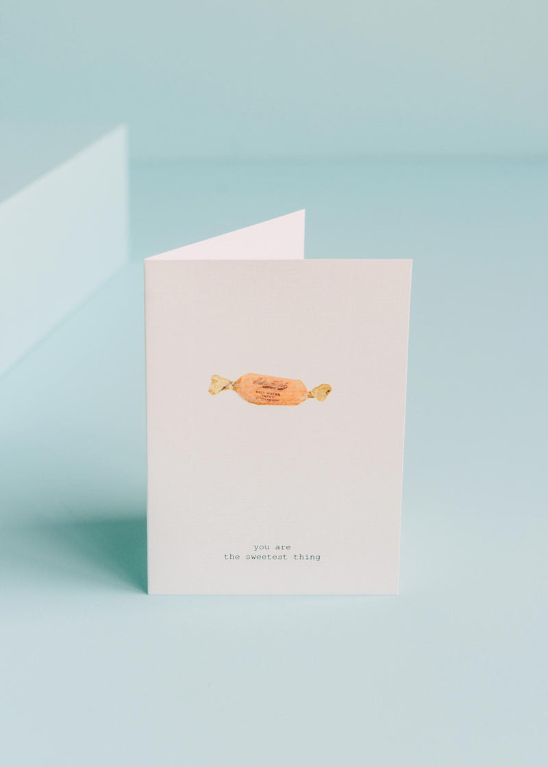 A TokyoMilk greeting card with a pastel blue background stands slightly open. The cover features an illustration of an orange candy with hand-glittered accents and the text "you are the sweetest thing" below by Margot Elena.