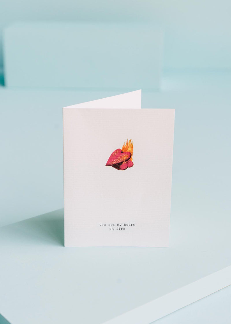 A TokyoMilk greeting card with a minimalist design, featuring an illustration of a flaming heart and the words "you set my heart on fire" printed on laid paper, standing against a soft blue background. Brand Name: Margot Elena