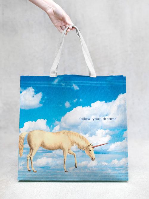 A hand holding a Margot Elena TokyoMilk Tote Bag - Unicorn Follow Your Dreams Market Tote with woven handles against a blue sky background and a white unicorn, alongside the inspirational text "follow your dreams" printed on it.